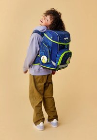 Blue backpack with neon green accents and geometric patterns, featuring multiple compartments and a circular tag. Worn with beige pants.