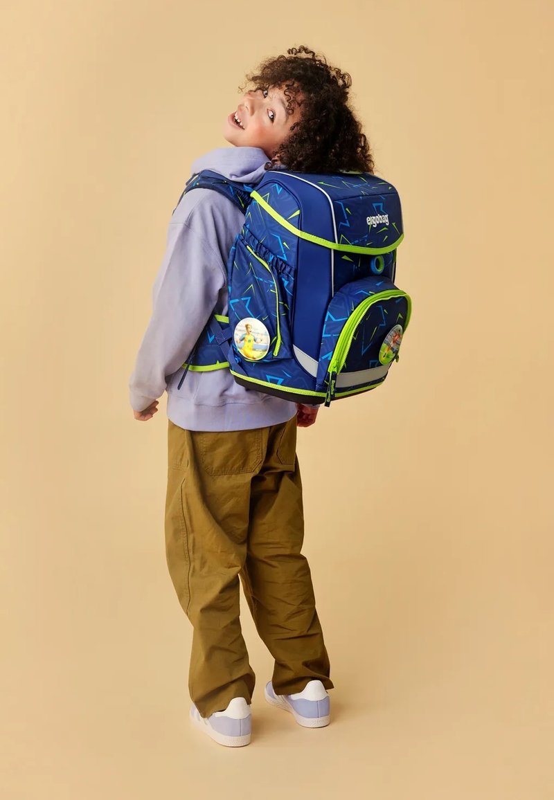 Blue backpack with neon green accents and geometric patterns, featuring multiple compartments and a circular tag. Worn with beige pants.