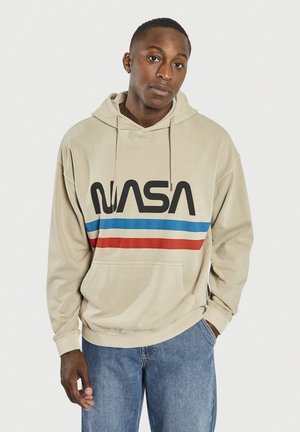Felpa con cappuccio beige caratterizzata da un grande logo "NASA" nero con strisce orizzontali rosse e blu sul petto. Il materiale sembra morbido.