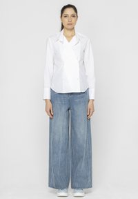 Biała koszula z podwójnym zapięciem i wyostrzonym kołnierzykiem, zestawiona z szerokimi, jasnoniebieskimi dżinsami i jasnoniebieskimi sneakersami. Proste, czyste linie.