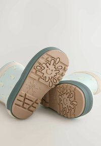 Blaue Gummistiefel mit beigen Akzenten und verspieltem Muster. Die strukturierten Sohlen zeigen ein lachendes Wasserspritzer-Design und tiefe Profile für guten Halt.