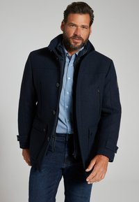 Marineblauer Wollmischungsmantel mit Knopfverschlüssen, zwei seitlichen Taschen und einer strukturierten Oberfläche. Das Innenfutter zeigt ein blaues Karomuster.