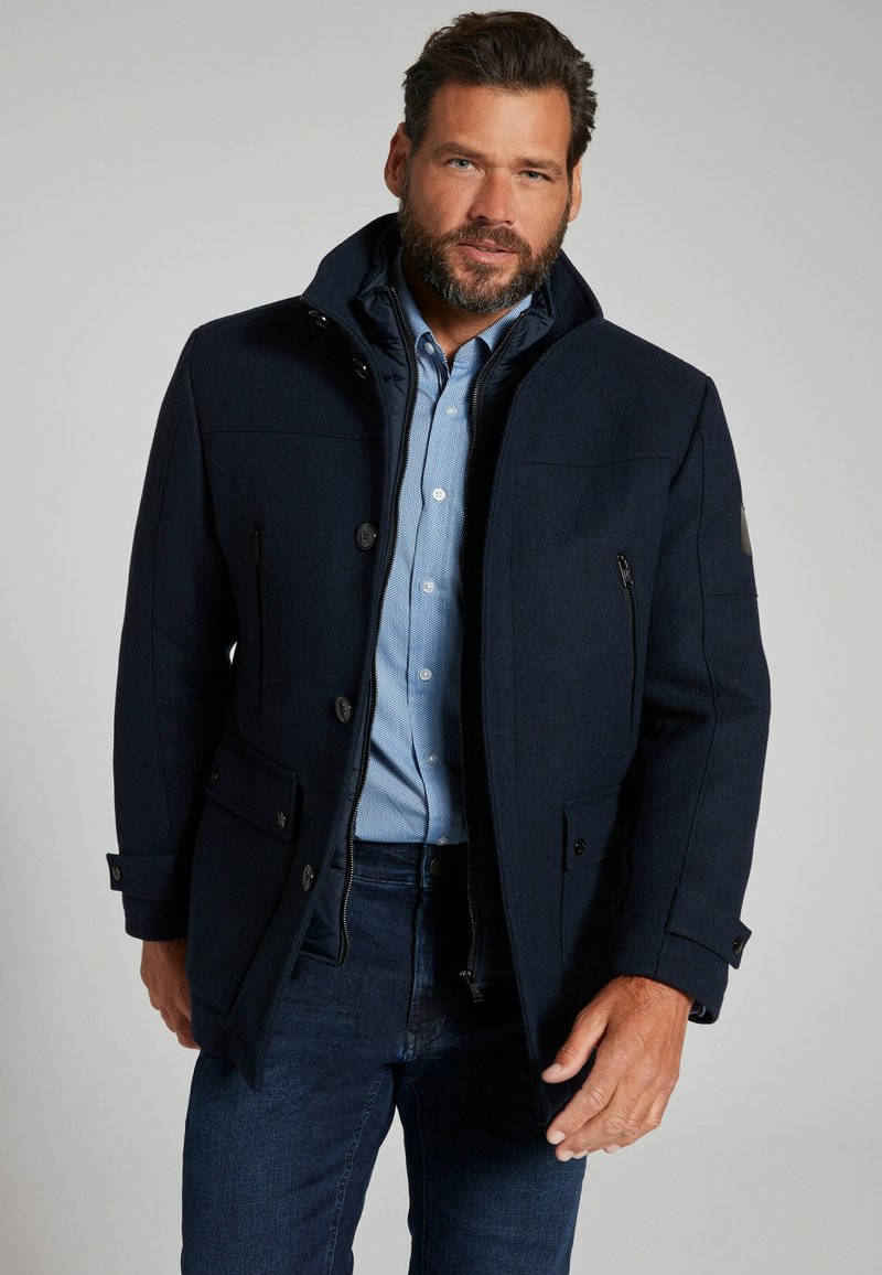 Marineblauer Wollmischungsmantel mit Knopfverschlüssen, zwei seitlichen Taschen und einer strukturierten Oberfläche. Das Innenfutter zeigt ein blaues Karomuster.