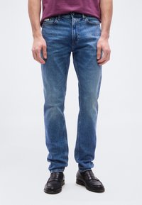 Άνδρας που φοράει μεσαίου μπλε στενά τζιν και μαύρα δερμάτινα μοκασίνια, στέκεται μπροστά σε απλό ανοιχτόφωτο φόντο.