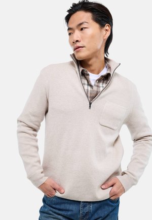 Homme avec les mains dans les poches portant un pull beige à fermeture éclair par-dessus une chemise à carreaux et un tee-shirt blanc, regardant sur le côté devant un fond uni.