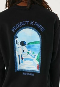 Sudadera negra con un gráfico vibrante inspirado en Santorini, con edificios blancos, cúpulas azules y acentos florales coloridos.