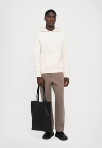 Kremet hettegensere med snor og frontlomme, kombinert med beige bukser. Svart totebag med lange håndtak og kraftige svarte sko.
