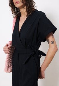 Zwarte wikkeljurk met korte mouwen en een strik in de taille. Kenmerkt zich door een gestructureerde stof. Houdt een roze geweven tas vast met een geometrisch patroon.