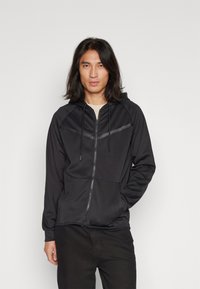 Sudadera con cremallera negra y capucha, hecha de una tela suave. Presenta acentos de rayas diagonales y bolsillos frontales. Costuras planas, corte relajado.