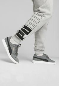 Szare sneakersy z czarnymi akcentami, wykonane z gładkiego materiału, o niskim profilu i białej podeszwie. W zestawie z szarymi spodniami dresowymi z czarnym logo.