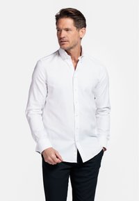 Chemise blanche à manches longues avec un col pointu et des poignets à boutons, fabriquée à partir d'un tissu texturé, associée à un pantalon sombre.