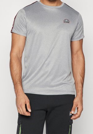 Mężczyzna ubrany w szarą koszulkę z krótkim rękawem z czerwoną taśmą na ramionach i czarne spodnie bojówki z zielonymi zamkami.