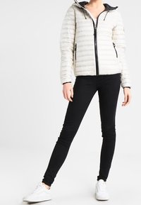 Bílá péřová bunda s horizontálním prošíváním, černým zipem a kapucí, spojená s černými skinny džíny a bílými teniskami.