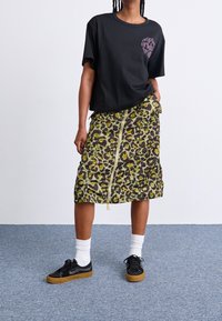 Camiseta gráfica negra, de corte relajado; falda de leopardo verde y marrón con textura fruncida y cremallera lateral; zapatillas negras con calcetines blancos.
