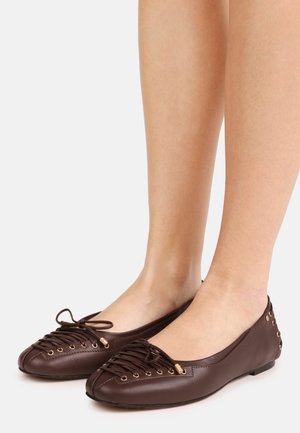 Brown leather ballet flats with crisscross laces, gold eyelets, bow detail, and stud accents worn on bare feet against a white background.