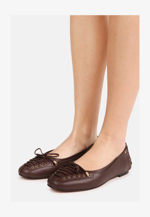 Bruine leren ballerina's met gekruiste veters, gouden veterogen, strikdetail en nagelaccenten, gedragen op blote voeten tegen een witte achtergrond.