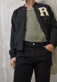 Chaqueta bomber negra con cremallera, que presenta un parche texturizado en blanco con una 'R', combinada con una camiseta verde y jeans negros. Tejido suave, ajuste informal.