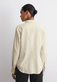 Cămașă lungă, de un bej deschis, cu mânecă lungă, dintr-un material moale și texturat, având un guler cu nasturi și o linie de tiv curbată. Design minimalist.