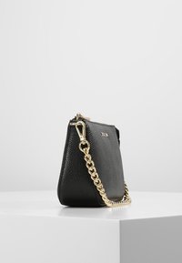 Bolso de mano de cuero negro con superficie texturizada, que presenta una correa de cadena dorada y una forma elegante y estructurada. Parte superior con cremallera y detalle del logo visible.