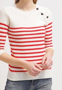 Pull en maille rayé rouge et blanc à manches courtes, avec un col rond et trois boutons décoratifs noirs sur l'épaule.