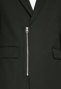 Schwarzer Blazer mit einem scharfen Design, ausgestattet mit einem auffälligen Metallreißverschluss vorne und zwei Seitentaschen. Strukturiertes Material.