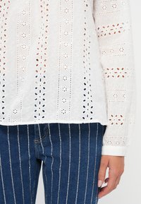 White embroidered blouse with cut-out patterns and long sleeves, paired with dark blue denim jeans featuring vertical metallic accents.