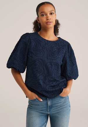 Haut brodé bleu marine avec des manches courtes bouffantes, comportant des motifs abstraits en relief et un col rond. Associé à un jean bleu clair.