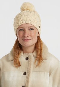 Gorro de punto crema con un patrón de trenzas texturizadas y un pom-pom esponjoso en la parte superior, combinado con una chaqueta sin cuello de color beige claro con un diseño de cuadros.