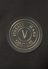 Černá kruhová nášivka loga Versace Jeans Couture se zlatým nápisem a centrálním "V" na černé látce.