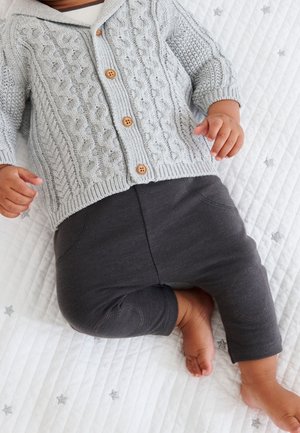 Bébé en cardigan gris tricot câblé avec des boutons en bois et un pantalon gris foncé, allongé sur une couverture matelassée blanche avec des étoiles argentées.