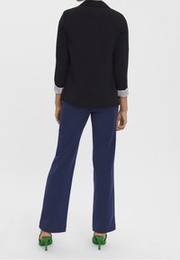 Černý blejzr s pruhovanými manžetami, spárovaný s námořnickými kalhotami a zelenými lodičkami na podpatku, pohled zezadu.