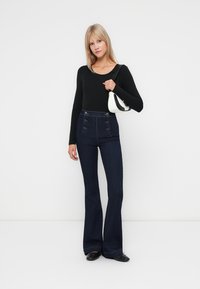 Haut noir à manches longues associé à un jean flare en denim foncé taille haute avec des accents de boutons, complété par un petit sac à main blanc.