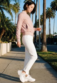 Young woman in light pink jacket and white pants standing on a sunny sidewalk with palm trees in the background.