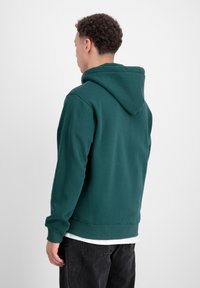 Felpa verde con cappuccio e vestibilità rilassata. Presenta polsini e orlo a costine. Il tessuto appare morbido con una texture liscia.