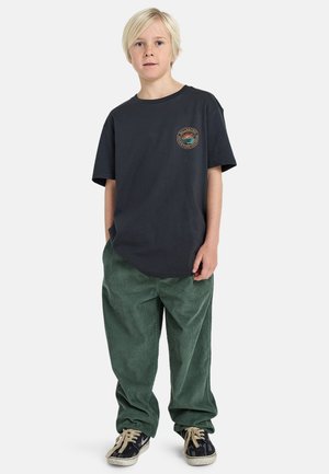 Maglietta nera in cotone con logo rotondo sul petto, abbinata a pantaloni in corduroy verdi e scarpe da skate nere.