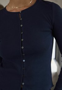 Tumed ribikangast nööpidega ülikond, millel on ümmargune kaelus, seitse esist nööpi ja veidi lühendatud hem, millel on õmbluse detail.