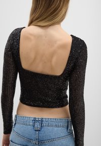 Schwarzes langärmliges Oberteil mit Pailletten und einem quadratischen Rückendesign. Verfügt über strukturierte Stoff, figurbetonten Schnitt und eine cropped Länge. Kombiniert mit hellblauen Jeans.