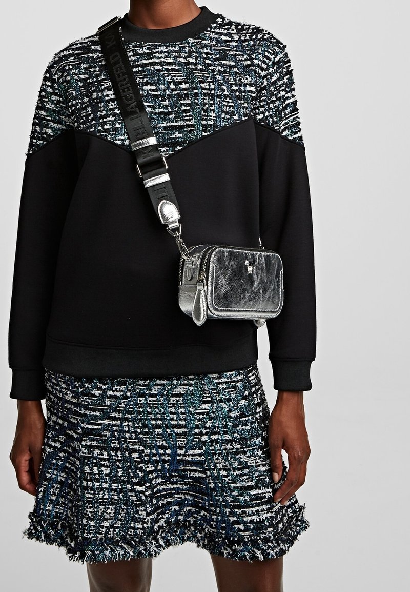 Set di maglione e gonna con texture nera e multicolore, abbinato a una borsa a tracolla metallica argentata, indossato da un modello su sfondo semplice.