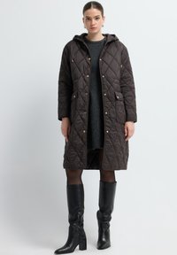 Gevulde, bruine lange jas met capuchon en drukknoopsluitingen, zijzakken, gedragen over een donkere trui, gecombineerd met kniehoge zwarte leren laarzen.