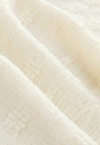 Tissu tricoté couleur crème avec un motif doux et texturé comprenant des points torsadés et des points boules en plis diagonaux.