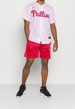 Biała baseballowa koszulka w cienkie prążki z logo "Phillies" w kolorze czerwonym, zestawiona z czerwonymi siateczkowymi szortami i czarnymi butami z zielonymi akcentami.