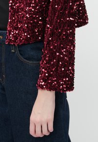 Burgundijas krāsas secinējuma jaka ar teksturētu virsmu un trim ceturtdaļām piedurknēm, apvienota ar tumšās džinsa biksēm, izceļot spīdumu un dizainu.