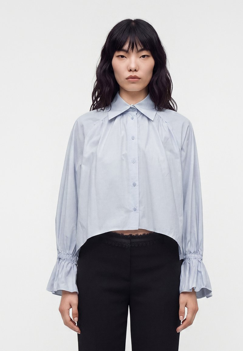 Femme aux longs cheveux noirs portant un chemisier bleu clair boutonné avec des manches volantées et un pantalon noir sur fond blanc uni.