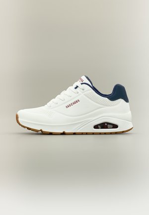 Valge Skechersi sportlik toss tumesinise kaelusega, nähtava õhavahtpõhjaga ja kummisest kollaka tallaga ühevärvilisel hallil taustal.