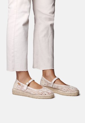 Stopy w beżowych szydełkowych espadrylach z paskami wokół kostek, w połączeniu z jasnymi, krótkimi spodniami.