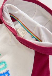 Megztinis su gobtuvu, bordo spalvos apvadai, kūno spalvos šviesus korpusas. Gobtuvo viduje yra vaivorykštės detalė ir žymė su užrašu „Little Bird by BODEN“.