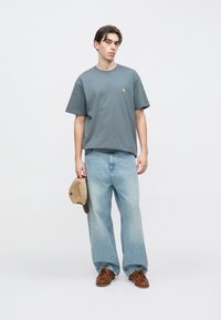 Νεαρός άνδρας με φαρδιά ανοιχτό μπλε τζιν παντελόνια, γκρι μπλούζα και καφέ παπούτσια, κρατώντας ένα μπεζ και καφέ καπέλο στο ένα χέρι,  στέκεται μπροστά σε λευκό φόντο.