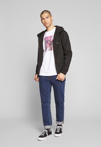 Sort letvægtsjakke med hætte, båret over en hvid t-shirt med et blomstermotiv, sammen med blå jeans og sorte sneakers.