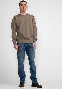 Bruine sweatshirt met ronde hals en geribbelde manchetten, gecombineerd met lichtblauwe jeans en donkere schoenen; heeft een kleine grafische afbeelding op de borst.