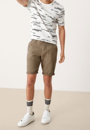 T-shirt blanche avec le texte noir "ANYTHING", shorts beiges roulés, baskets blanches et chaussettes rayées noir et blanc. Tenue simple et décontractée.